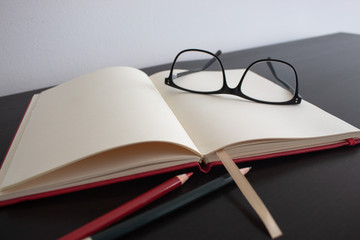 Black glassess with red color pencil and green pencil on blank red notebook and wooden table.