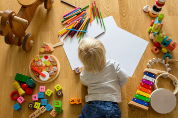 Little blonde toddler boy, drawing with pastels and coloring pens, playing with wooden toys