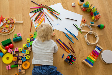 Little blonde toddler boy, drawing with pastels and coloring pens, playing with wooden toys