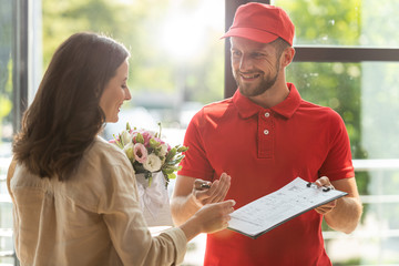bearded and happy delivery man holding clipboard and pen near beautiful woman