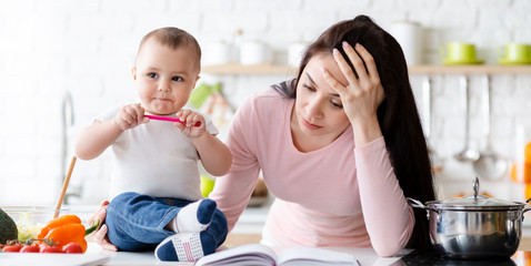 Cute baby biting pen, tired mom sitting aside, kitchen interior