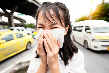 Portrait closeup of child girl blowing nose in paper handkerchief,asian woman sneezing in a tissue in the city street,toxic fumes from car,bad smell,concept of air pollution,dust allergies,health care