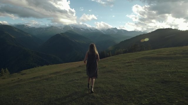 Camera follows hipster millennial young woman in a long dress developing in the wind running up on top of mountain summit at sunset, raises arms into air, happy and drunk on life, youth and happiness.