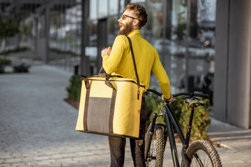 Young courier delivering food in a yellow thermo bag, walking with bicycle on the street in the city. Delivery service concept