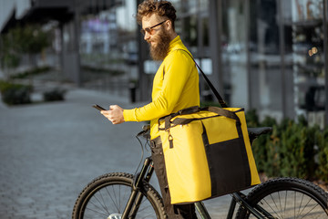 Young bearded courier delivering food in thermal bag on a bicycle, checking order with a smart phone while standing on the street outdoors