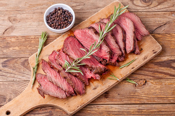 Slices of medium rare roast beef meat on wooden cutting board, pepper and rosemary branches on...