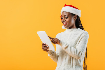 African American woman in winter sweater and Santa hat tapping on screen of digital tablet isolated on yellow
