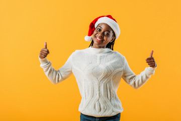 African American woman in winter sweater and Santa hat showing like signs isolated on yellow