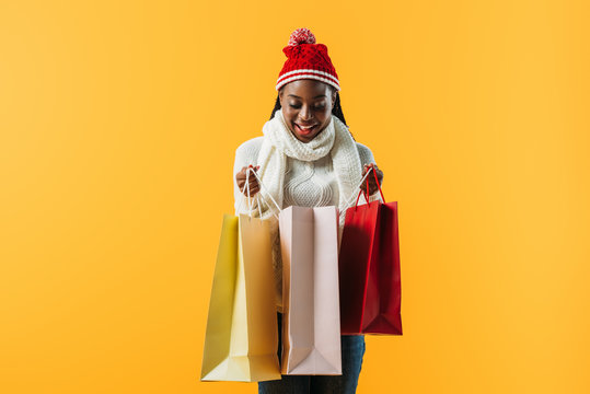 African American woman in winter outfit holding shopping bags and looking inside isolated on yellow