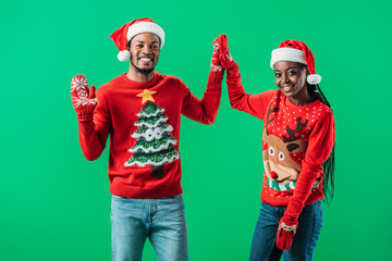 African American couple in red Christmas sweaters and Santa hats giving high five to each other and looking at camera isolated on green