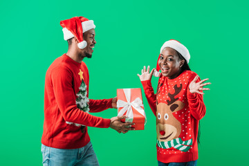 African American man in red Christmas sweater and Santa hat giving gift box to surprised woman isolated on green