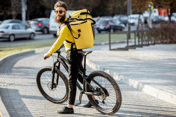 Naklejka na ściany i meble Portrait of a young courier delivering food with a yellow thermal backpack on a bicycle in the city. Food delivery service concept