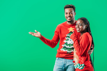 African American man pointing with hand and look at camera near woman in Christmas sweater isolated on green