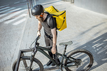 Male courier taking a bicycle from the parking outdoors, delivering food with yellow thermal...