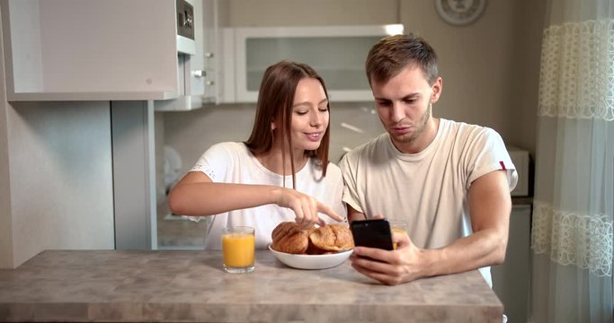 Young lovers in white home t-shirts eating croissants, drinking juice and surfing internet using smartphone in kitchen