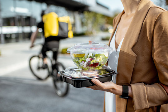 Businesswoman holding lunchboxes with fresh takaway food outdoors. Male courier on a bicycle on the background. Takeaway restaurant food delivery concept