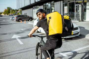 Young bearded courier delivering food with a yellow thermal backpack, riding a bicycle in the city. Food delivery service concept - obrazy, fototapety, plakaty