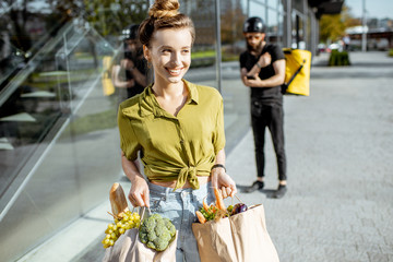 Young and happy woman carrying bags full of fresh food received from a delivery man. Courier standing with delivery bag on the background