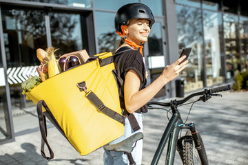 Young female courier delivering fresh food, standing with smartphone and backpack full of products...
