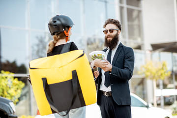 Young female courier delivering takeaway lunches with a thermal bag to an office worker dressed in the suit near the office building outdoors