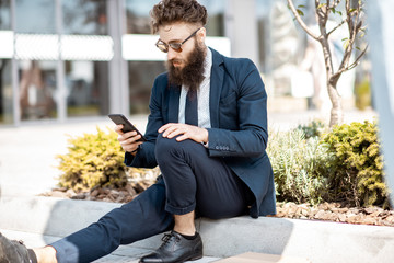 Portrait of an elegant bearded businessman dressed in the suit sitting with phone near the office building outdooors