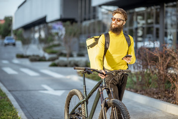 Young bearded courier delivering food on a bicycle, checking order with a smart phone while...