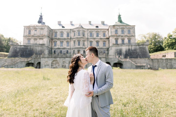 Fabulous Asian wedding couple posing in front of an old medieval castle, hugging and kissing on a sunny day.