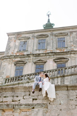 Fototapeta na wymiar Beautiful Asian wedding couple poses while sitting on the stone wall of an old ruined castle