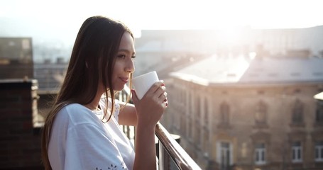 Attractive young lady drinking hot coffee early in morning standing on balcony and looking at street, relaxation