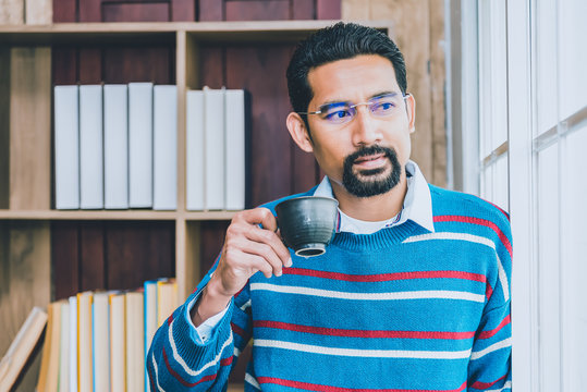 Arab businesman in casual clothes taking a break from work, standing near the window holding cup of coffee blurred black ground of bookcase.