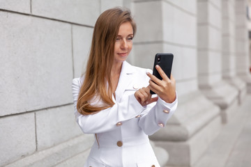 Happy young businesswoman in formal wear holding and touching the smartphone near the office building. Corporate lifestyle.