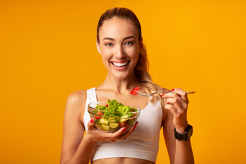 Fitness Girl Eating Vegetable Salad Standing Over Yellow Background - obrazy, fototapety, plakaty