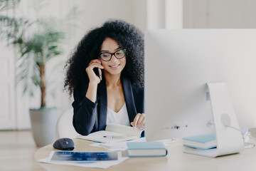 Obraz premium Photo of satisfied Afro American woman calls via smartphone, makes notes in notepad, involved in working process, sits at desk with computer, wears spectacles, formal clothes, has phone communication