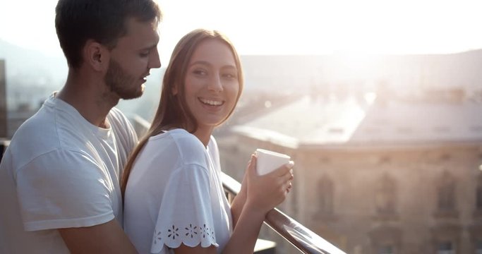 Attractive young man embracing pretty girlfriend drinking coffee on balcony early in morning, dating