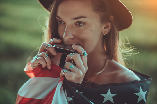 Portrait of boho chic woman in hat with american flag wearing silver rings with turquoise stone playes on harmonica outdoors. Jewelry indie girl with hippie style and boho fashion. Travel to america