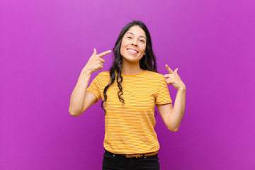 young pretty latin woman smiling confidently pointing to own broad smile, positive, relaxed,...