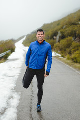Athlete warming up and stretching legs before running under the snow on winter mountain road.