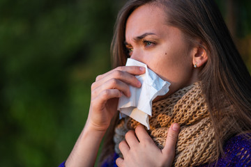 Sick unhappy woman in knitted scarf caught a cold in the fall and suffers from stuffy, runny nose and uses a paper napkin during sneezing outdoors. Seasonal colds and infections. Autumn cold and flu