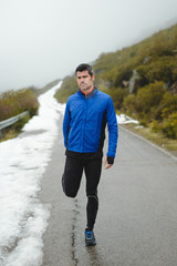 Athlete warming up and stretching legs before running under the snow on winter mountain road.