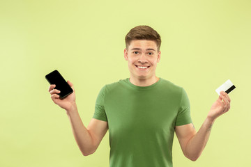 Caucasian young man's half-length portrait on green studio background. Beautiful male model in shirt. Concept of human emotions, facial expression, sales, ad. Holding phone and card, online payments.