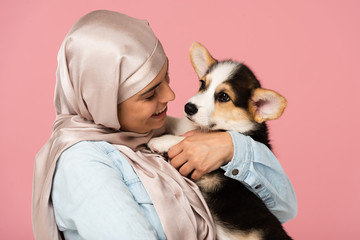 beautiful muslim girl in hijab holding Welsh Corgi puppy, isolated on pink