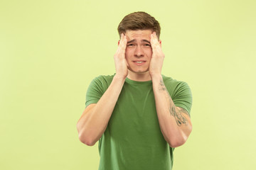 Caucasian young man's half-length portrait on green studio background. Beautiful male model in shirt. Concept of human emotions, facial expression, sales, ad. Suffering from headache, healthcare.