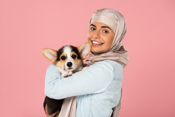 smiling muslim woman in hijab holding Welsh Corgi puppy, isolated on pink