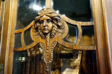 Fototapeta na wymiar Wooden door of an antique bookcase (closet) with a carved woman face. Close-up photo. Vintage furniture sculpture detail. European museum. Old books
