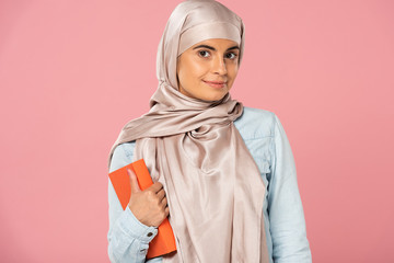 attractive muslim student in hijab holding book isolated on pink