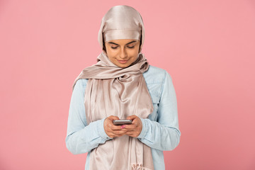 beautiful smiling muslim girl using smartphone isolated on pink