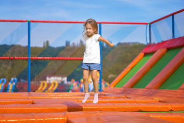 Fototapeta na wymiar Little pretty girl having fun outdoor. Jumping on trampoline in children zone. Happy girl jumping on the yellow trampoline in Amusement park