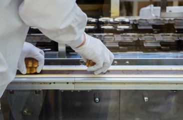 Man in white gloves is taking a cookie. This is a factory for dessert production. White uniform.