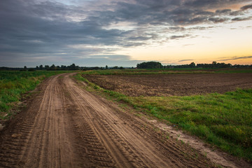 Fototapeta na wymiar Brown dirt road through fields and clouds after sunset