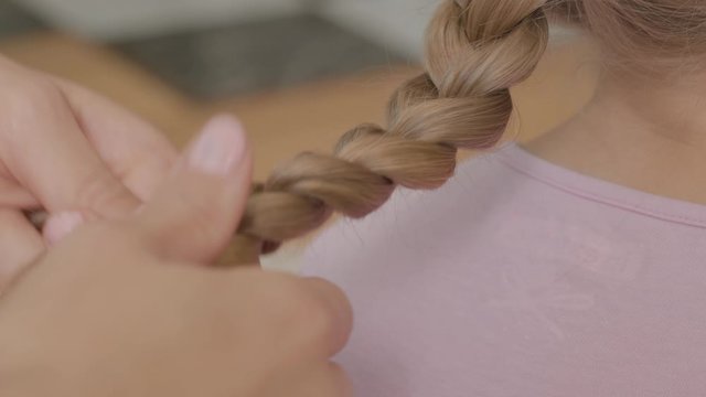Close-up hands of young woman braids a braid of her blond daughter. Concept of motherhood, childhood, one parent. Leisure indoors. Happy family